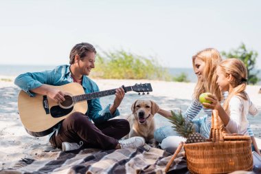 Akustik gitar çalan adamın seçici odağı aile ve Golden Retriever plajda 