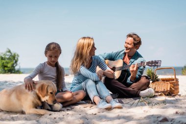 Kocasına bakan kadının seçici odak noktası akustik gitar çalması. Kızının yanında Golden Retriever ile plajda. 