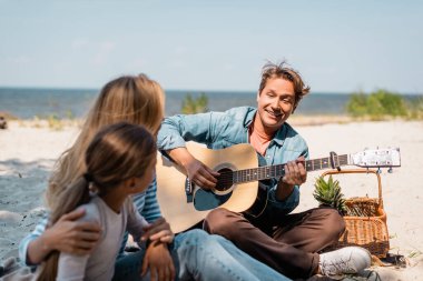 Sahilde eş ve çocuğun yanında akustik gitar çalan adamın seçici odak noktası. 