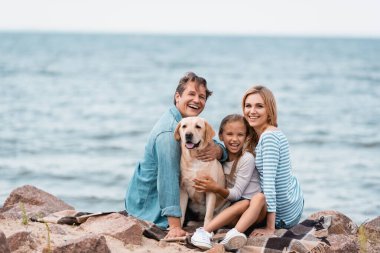 Ailenin seçici odak noktası deniz kenarında Golden Retriever 'ı okşarken kameraya bakmak. 