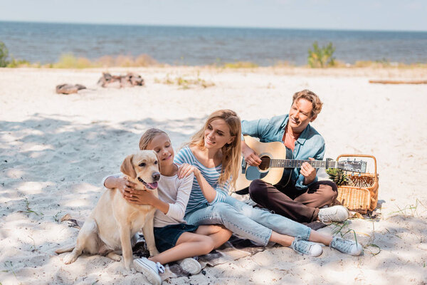 Ребенок обнимает золотистого ретривера рядом с матерью и отцом, играя на акустической гитаре на пляже 