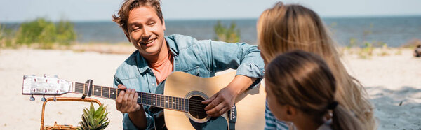 Horizontal image of man playing acoustic guitar near family on beach 