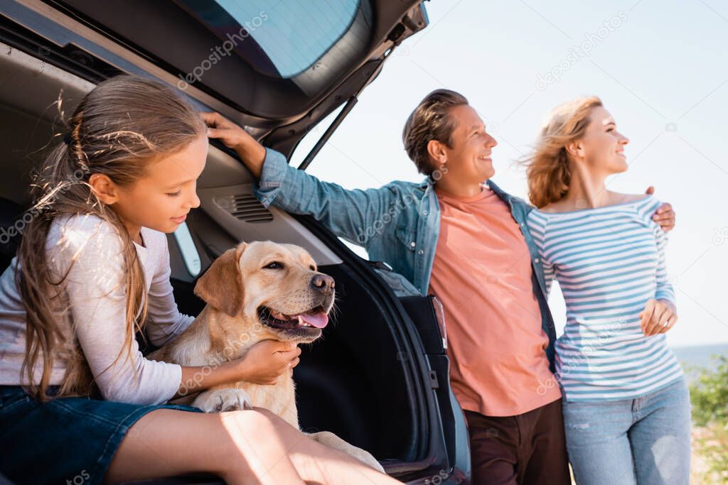 Selective focus of child and golden retriever sitting in car trunk near parents outdoors 