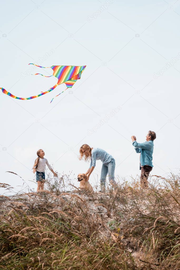 Selective focus of man holding kite near wife and daughter with golden retriever on grassy hill
