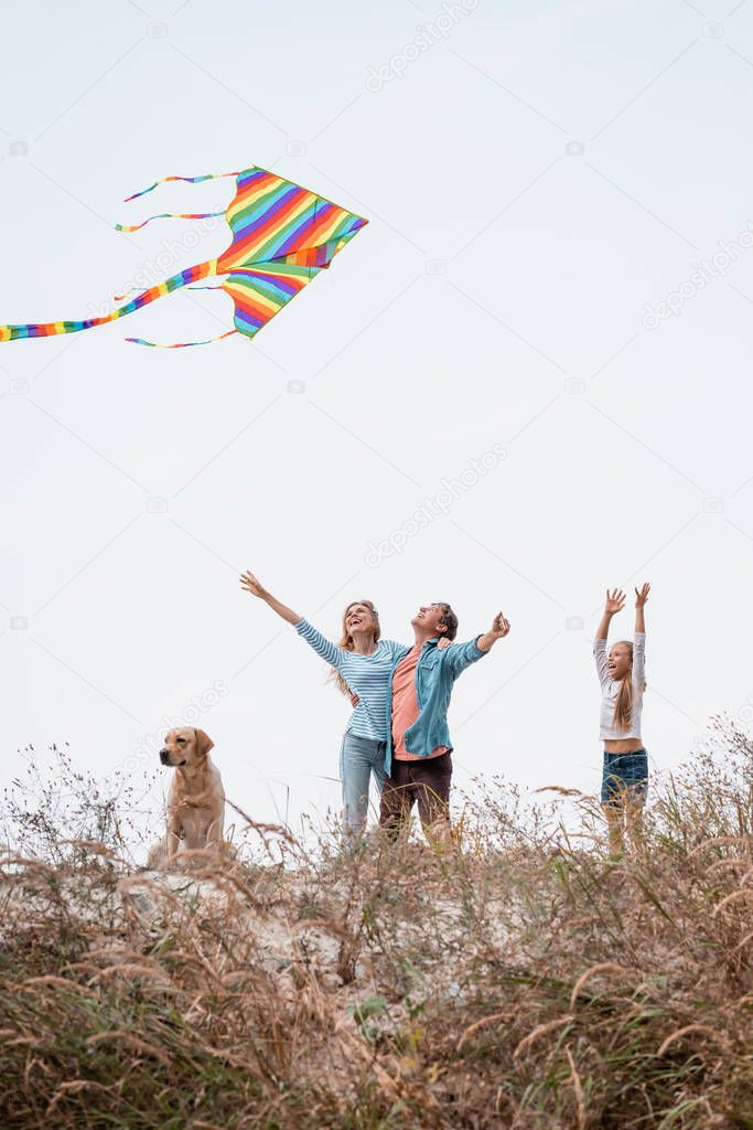 Selective focus of parents with kite hugging near daughter and golden retriever on grassy hill