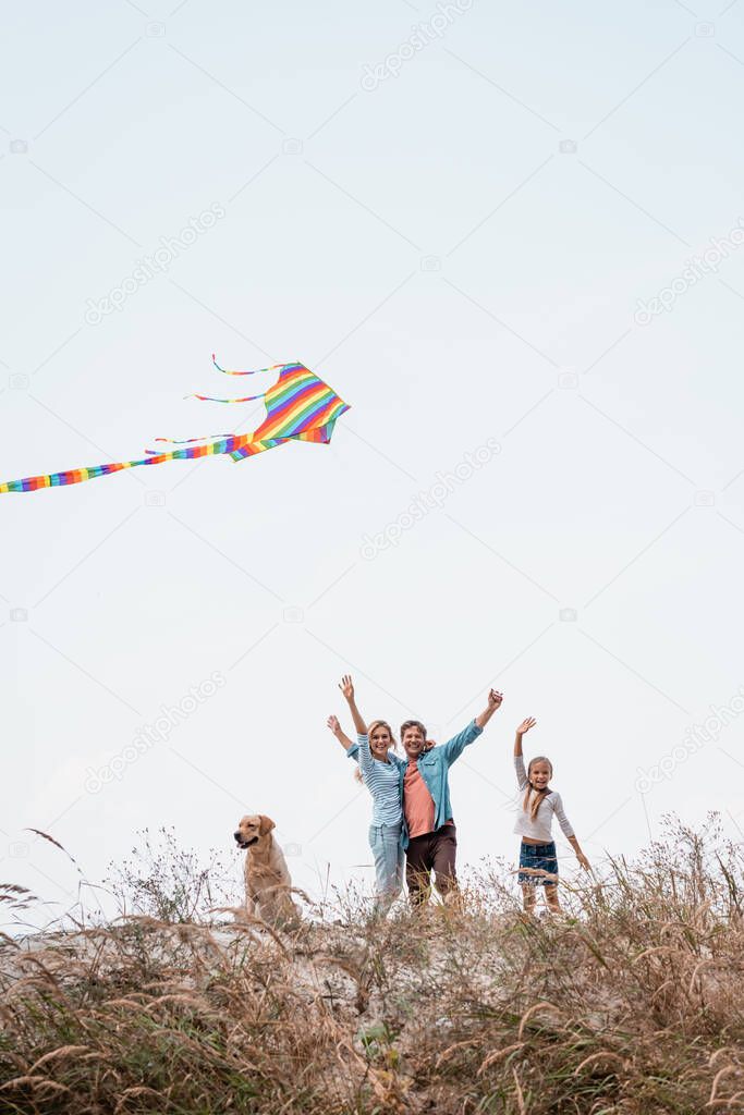 Selective focus of family with golden retriever and kite waving at camera on grassy hill 