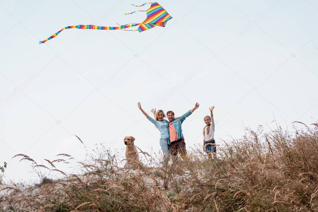 Selective focus of family with kite and golden retriever waving hands at camera on hill during weekend 
