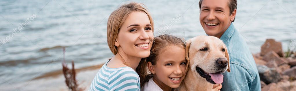Panoramic shot of parents and daughter with golden retriever looking at camera on beach