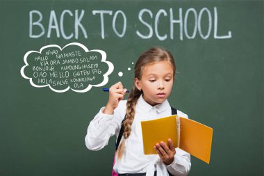 concentrated schoolgirl holding copy book and pen near chalkboard and greeting lettering in classroom  clipart