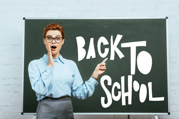 shocked teacher touching face and pointing with finger at chalkboard with back to school lettering in classroom