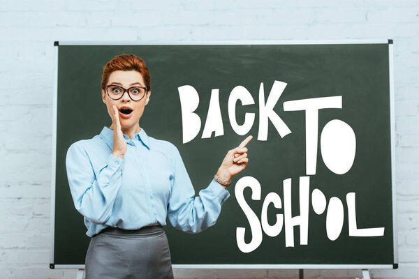 shocked teacher touching face and pointing with finger at chalkboard with back to school lettering in classroom 