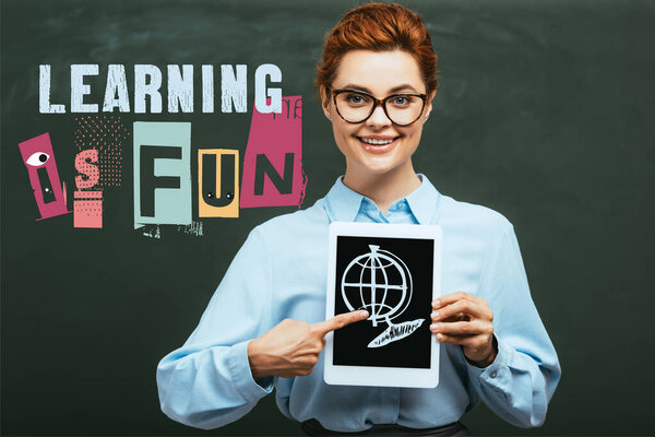 teacher pointing with finger at digital tablet with blank screen and globe illustration near chalkboard with learning is fun lettering 