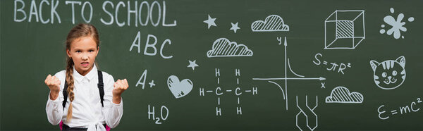 horizontal image of angry schoolgirl showing clenched fists near chalkboard with back to school lettering and geometry formulas in classroom 