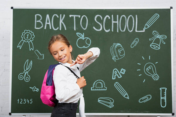 schoolgirl pointing with finger at backpack near chalkboard with back to school lettering and illustration 