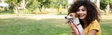 Genç bir kadının panoramik görüntüsü. Kameraya bakıyor ve köpeği tutuyor.