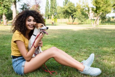 Seçici odak noktası, genç bir kadının köpekle çimenlerde oturup kameraya bakması.