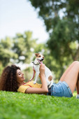 Fröhliche Frau im Sommer-Outfit liegt mit Jack Russell Terrier-Hund auf grünem Rasen