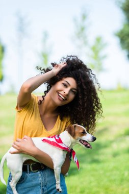 Neşeli, esmer kadın kıvırcık saçlara dokunurken Jack Russell Terrier köpeğini tutuyor.