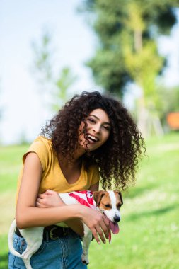 Kıvırcık kadın kameraya bakıyor ve Jack Russell Terrier köpeğini tutarken gülüyor.