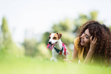 Kıvırcık kadının Jack Russell Terrier köpeğiyle çimenlikte dinlenirken gülüşünün seçici odağı.