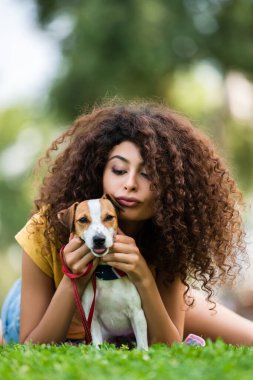 Esmer, kıvırcık saçlı kadın yeşil çimlerin üzerinde Jack Russell Terrier köpeğiyle rahatlarken dudaklarını büzüyor.