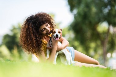 Jack Russell Terrier köpeğine sarılıp çimenlikte uzanan neşeli kadının seçici odağı.