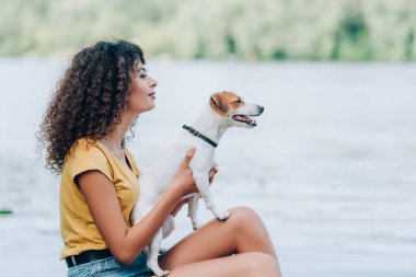 Yazlık kıyafetli kıvırcık kadın, Jack Russell Terrier köpeği tutuyor ve göl kenarında otururken gözlerini kaçırıyor.