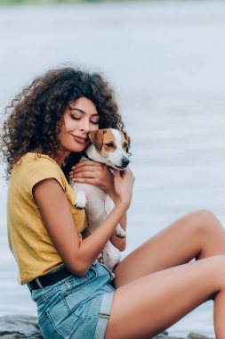 Yazlık kıyafetli genç bir kadın göl kenarında gözleri kapalı Jack Russell Terrier köpeğine sarılıyor.