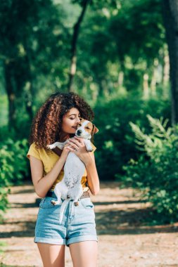 Yazlık kıyafetli genç, kıvırcık kadın parkta Jack Russell Terrier 'ı öpüyor.