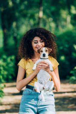 Yazlık kıyafetli neşeli kadın parkta Jack Russell Terrier köpeği tutuyor.