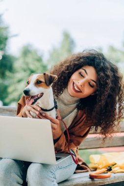 Keyifli serbest yazar dizüstü bilgisayarla bankta otururken Jack Russell Terrier köpeğine sarılıyor.