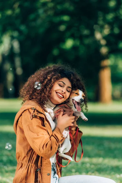 Yağmurluklu kadının seçici odağı parkta sabun köpüklerinin yanında Jack Russell Terrier 'ı kucaklıyor.