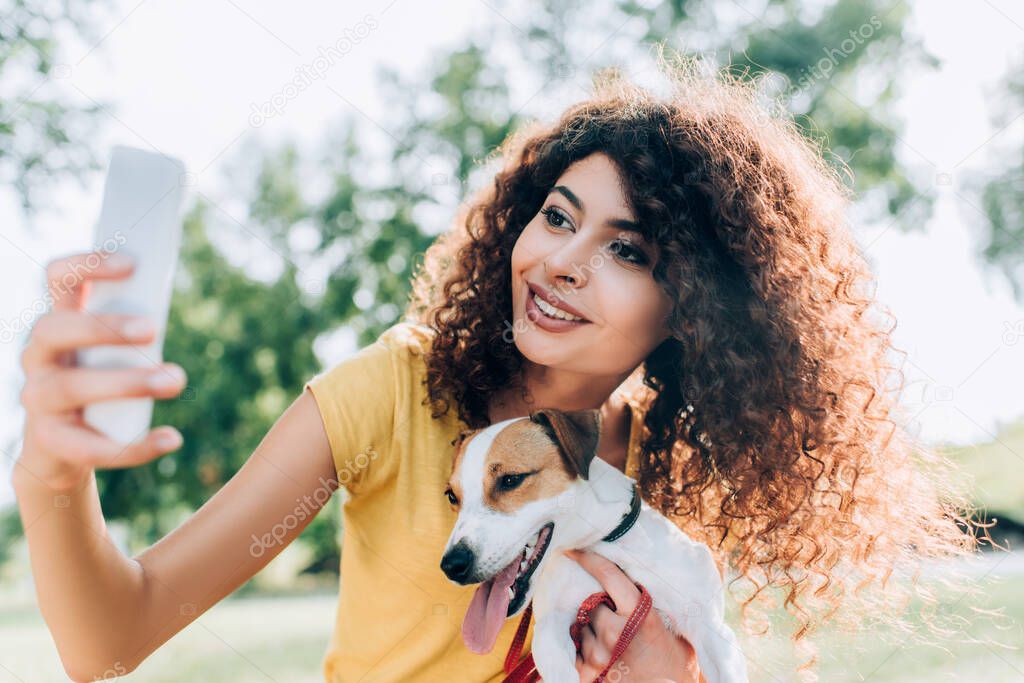 young, curly woman cuddling jack russell terrier dog while taking selfie on cellphone in park