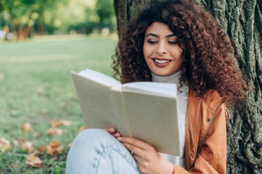 Selective focus of curly woman in autumn outfit reading book in park  clipart