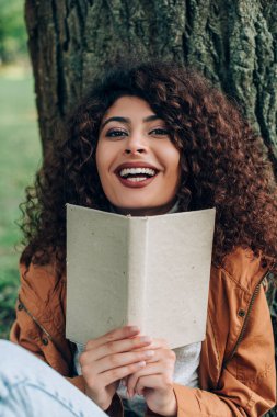 Kitaplı kadının seçmeli odak noktası parktaki ağacın yanındaki kameraya bakması. 