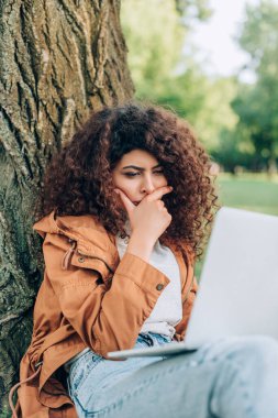 Kotlu ve yağmurluklu dalgın serbest çalışanın seçmeli odak noktası parktaki ağacın yanındaki dizüstü bilgisayara bakıyor. 
