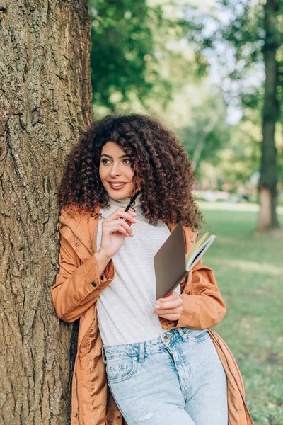 Mujer Rizada Impermeable Sosteniendo Pluma Cuaderno Cerca Del Árbol Parque —  Fotos de Stock