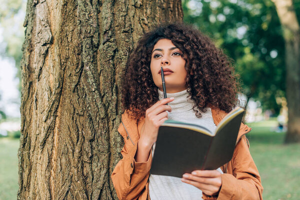 Selective focus of dreamy woman in raincoat holding pen and notebook near tree 