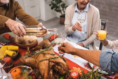 cropped view of senior man pouring white wine during thanksgiving dinner with multicultural family clipart