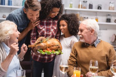 Seçici odak noktası, Afrika kökenli Amerikalı bir kadının hindiyi kızının ve sofrada çok ırklı bir ailenin tepsisinde tutması. 