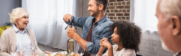 多文化家族で感謝祭の夕食の間に白ワインの瓶を開ける男のパノラマの概念 — ストック写真