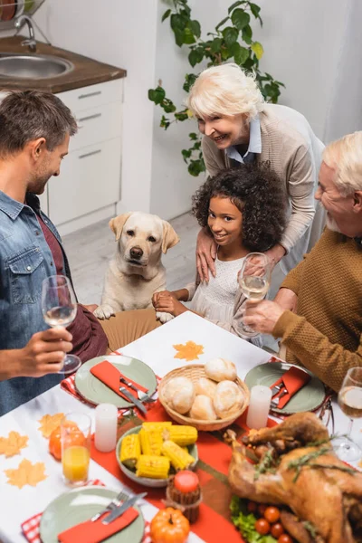 multicultural family and golden retriever near table served with festive thanksgiving dinner