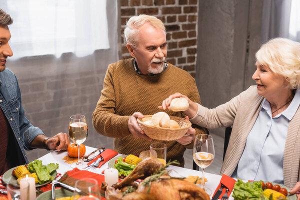 senior woman taking bun from wicker basket near husband and son during thanksgiving dinner