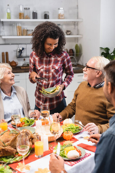 selective focus of joyful african american woman serving vegetables during thanksgiving dinner with multicultural family