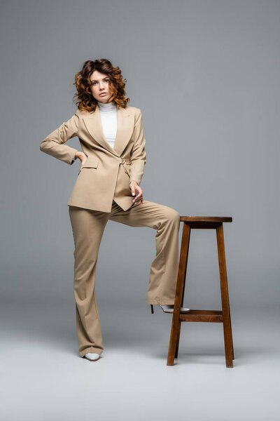 элегантная женщина в бежевом костюме позирует возле деревянного стула на сером
