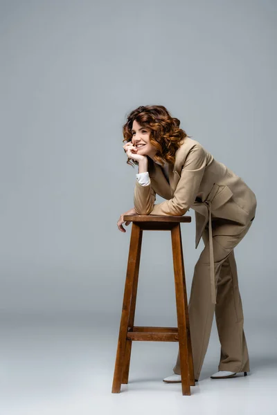 Elegantní Žena Béžovém Obleku Pózuje Dřevěné Židli Usmívá Šedou — Stock fotografie