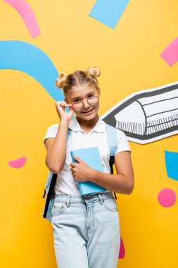 Sarı arka plan üzerine kağıt kalem ve çok renkli elementlerle kitap tutarken gözlüklere dokunan neşeli öğrenci.