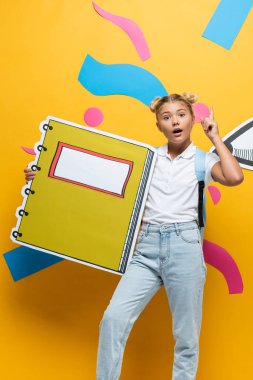 Renkli elemanlar ve kağıt kesikli kalemle sarı arka planda fikir jesti sergileyen okul kızı.