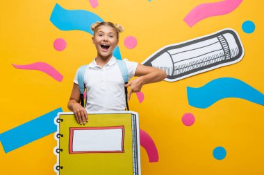 Sarı arka planda, kağıt kesiği kalem ve dekoratif elementlerle dizüstü bilgisayarı işaret eden hayrete düşmüş okul çocuğu. 