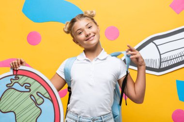 Kağıt kalem ve renkli elementlerle sarı arka planda küresel maketi tutan öğrenci memnun.